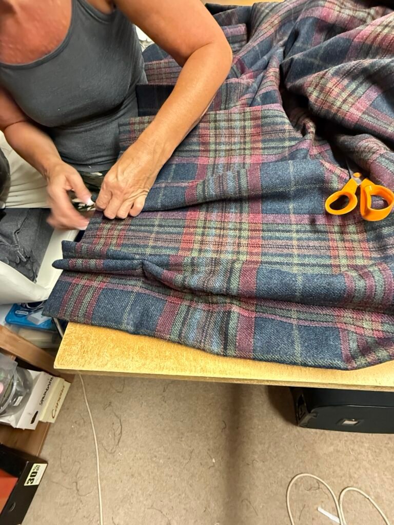 A close-up showing the corner of a lined tartan curtain being carefully sewn by hand. Hand stitching allows the fabric and lining to be secured neatly while keeping visible stitches to a minimum.
